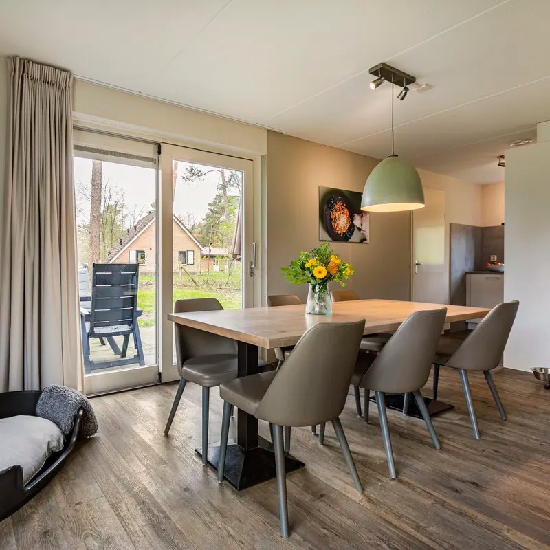Landal Twenhaarsveld: Een moderne woonruimte met een grote eettafel en leren stoelen, verlicht door een hanglamp. Naast de tafel staat een vaas met bloemen en een hondenbed is zichtbaar aan de linkerkant van de afbeelding.