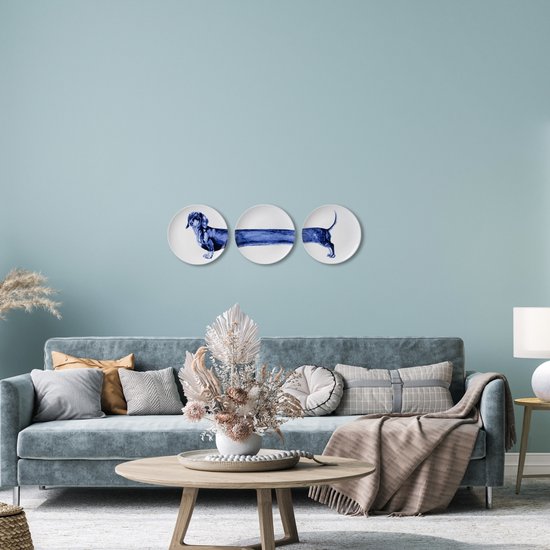 Een creatieve wanddecoratie bestaande uit drie borden met een teckelmotief, op een muur boven een moderne bank met kussens en een decoratieve tafel.