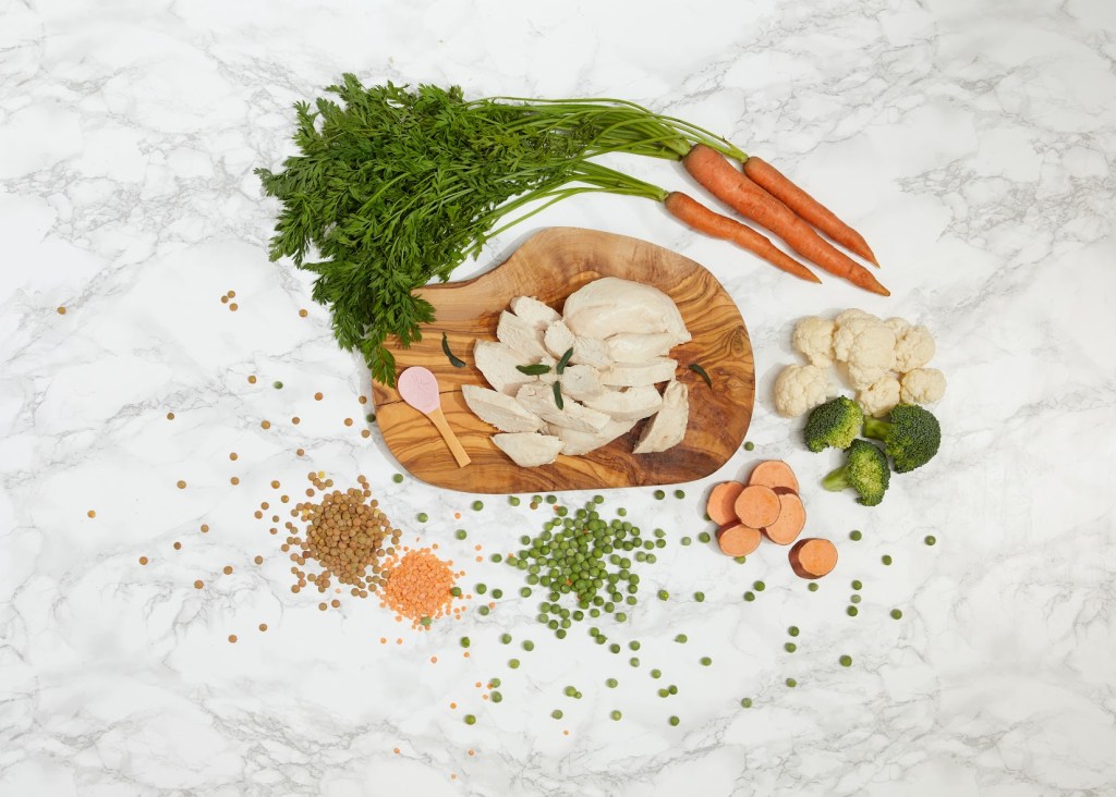 Een houten plank met gesneden kipfilet en verse groenten zoals wortelen, broccoli, bloemkool en zoete aardappel, omringd door linzen en groene erwten, op een marmerachtig oppervlak.